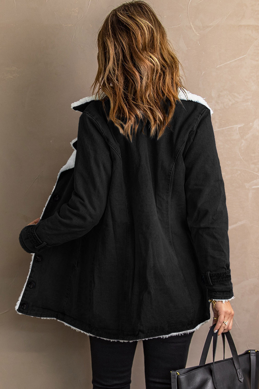 Donna di spalle con cappotto nero invernale foderato di pelliccia sintetica, capelli castani ondulati, borsa nera. Moda autunno inverno elegante.