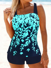 Donna in costume da bagno tankini blu con stampa foglie verdi, su sfondo spiaggia. Moda mare estiva, abbigliamento donna, look casual elegante.