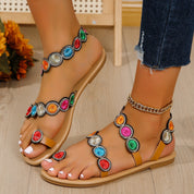 Sandali estivi da donna con cinturini decorati da pietre colorate, suola piatta beige, indossati con jeans. Accessorio moda casual e vivace.