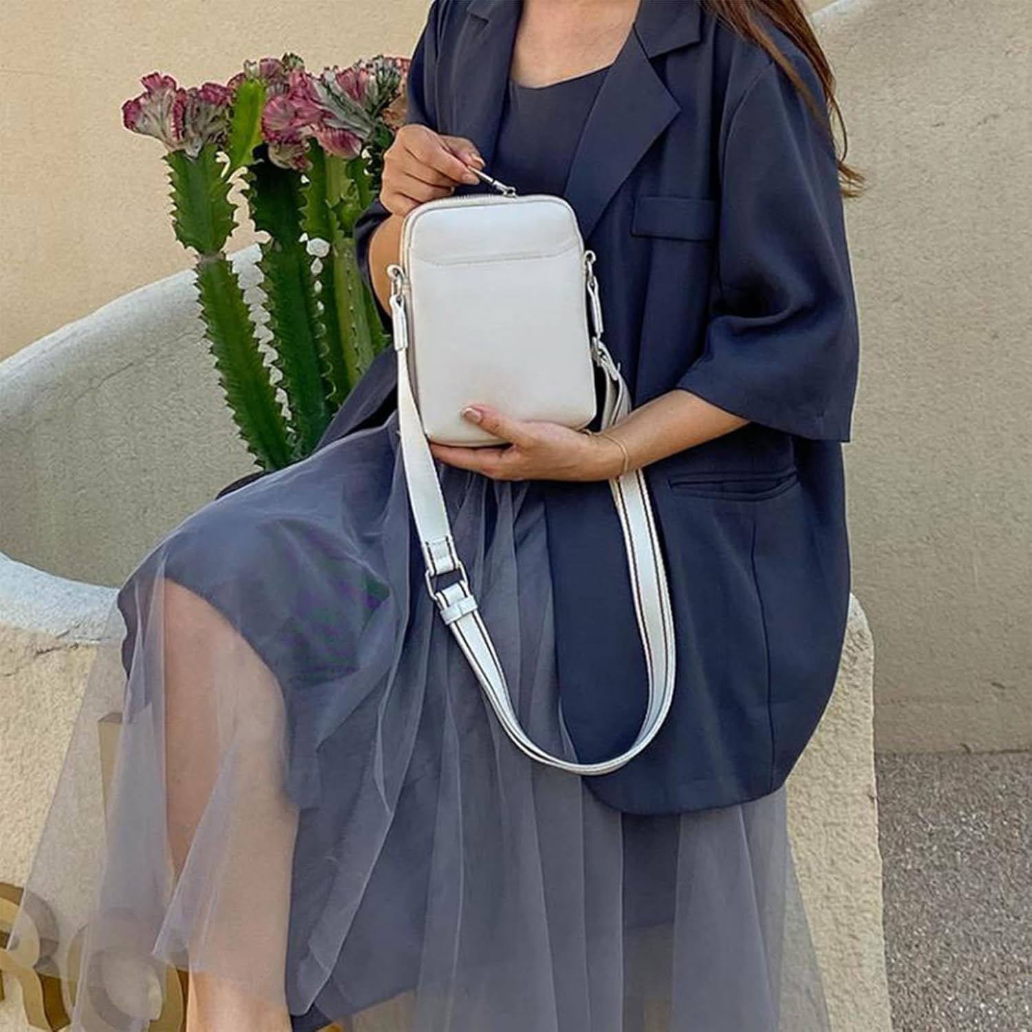 Donna seduta con abito blu e giacca, tiene una borsa a tracolla bianca. Sfondo con cactus. Moda elegante, accessori femminili, stile urbano.