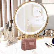 Specchio da trucco rotondo con cornice dorata su tavolo da toeletta, circondato da pennelli cosmetici e pochette rosa, in bagno moderno.