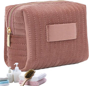 Astuccio cosmetico rosa trapuntato con cerniera dorata, accompagnato da asciugamani, spazzola e flaconi. Accessorio da viaggio elegante e pratico.