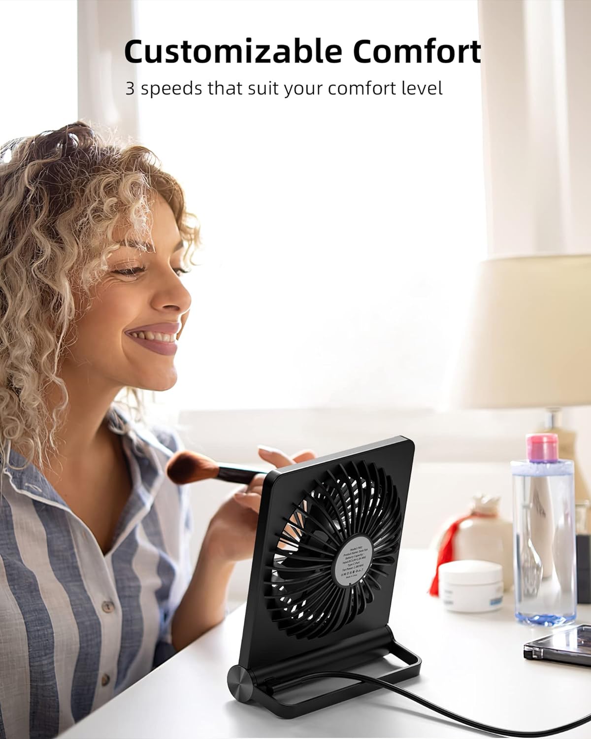 Donna sorridente usa ventilatore da tavolo nero con tre velocità, ideale per comfort personalizzato. Accessori trucco e bottiglia d'acqua sullo sfondo.