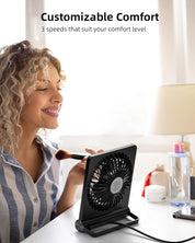 Donna sorridente usa ventilatore da tavolo nero con tre velocità, ideale per comfort personalizzato. Accessori trucco e bottiglia d'acqua sullo sfondo.