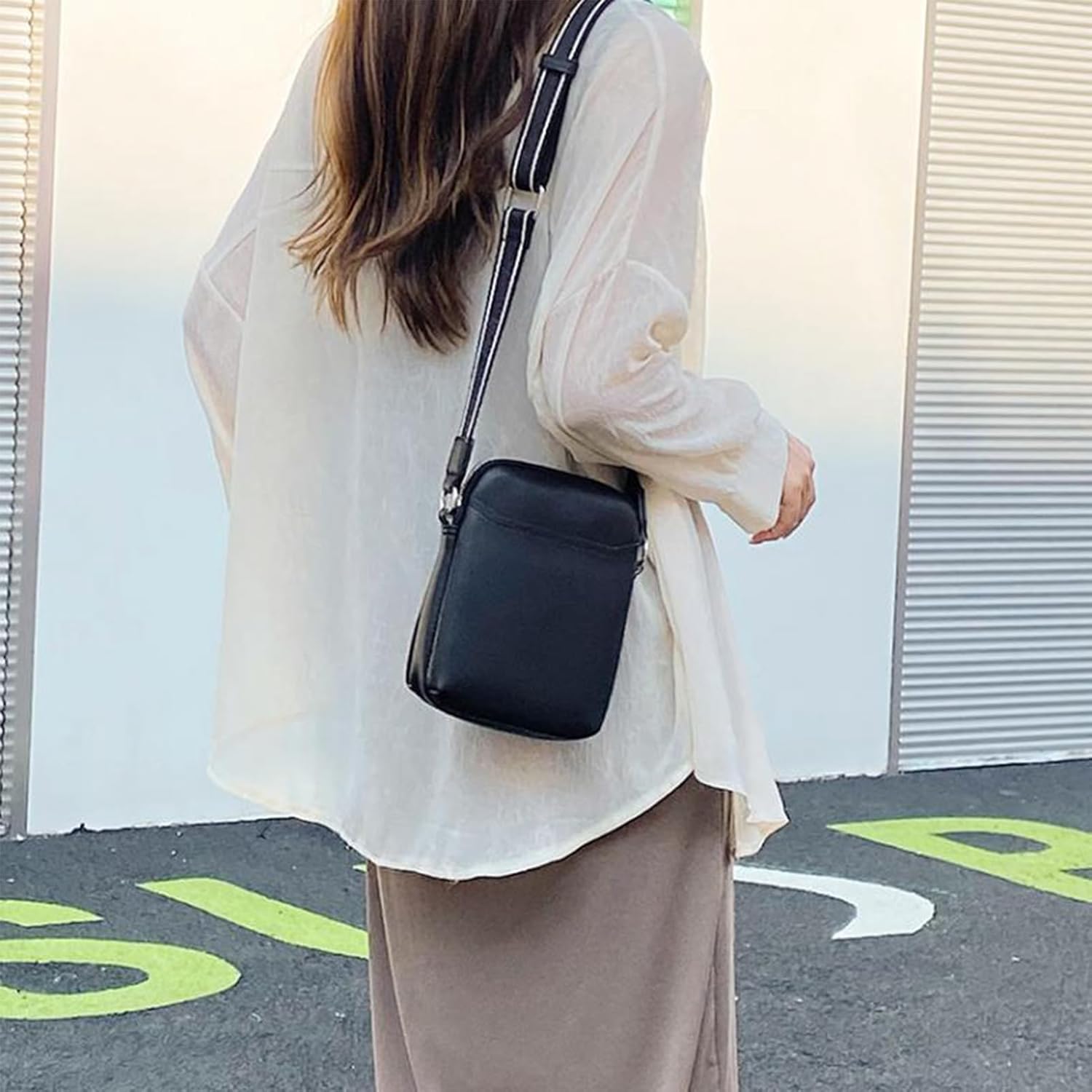 Donna con borsa a tracolla nera in pelle, indossa camicia bianca trasparente e gonna beige. Moda casual femminile, accessori eleganti.