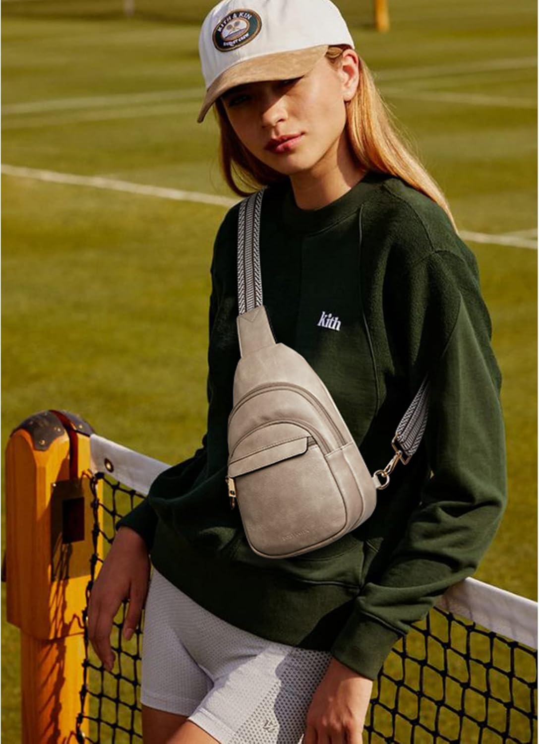 Giovane donna con cappellino bianco, felpa verde e marsupio beige, appoggiata a una rete da tennis. Moda casual sportiva all'aperto.