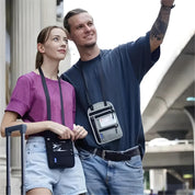 Due persone sorridenti con portadocumenti a tracolla, in viaggio. Sfondo urbano con valigia. Accessori pratici per viaggiatori moderni.