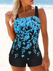 Donna in costume da bagno tankini nero e blu con motivo floreale, spalline larghe, su spiaggia. Moda mare, abbigliamento estivo, costumi da bagno donna.