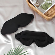 Maschera per dormire nera su tessuto di seta, con cinturino regolabile. Accessorio per il sonno, relax e viaggio. Cesto con fiori e libro accanto.