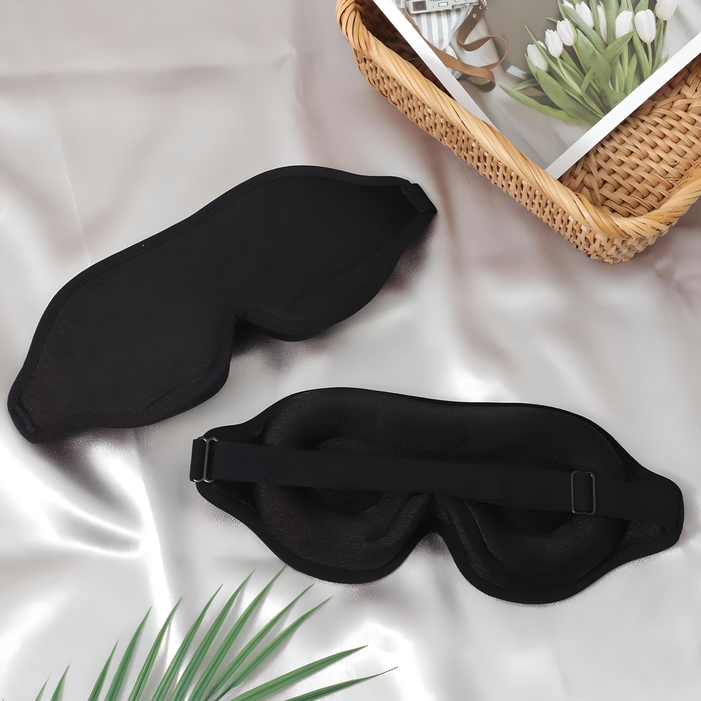 Maschera per dormire nera su tessuto di seta, con cinturino regolabile. Accessorio per il sonno, relax e viaggio. Cesto con fiori e libro accanto.