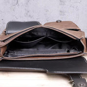 Borsa a tracolla in pelle marrone aperta, con scomparti interni neri e cuciture bianche, su sfondo grigio. Elegante e funzionale per uomo e donna.