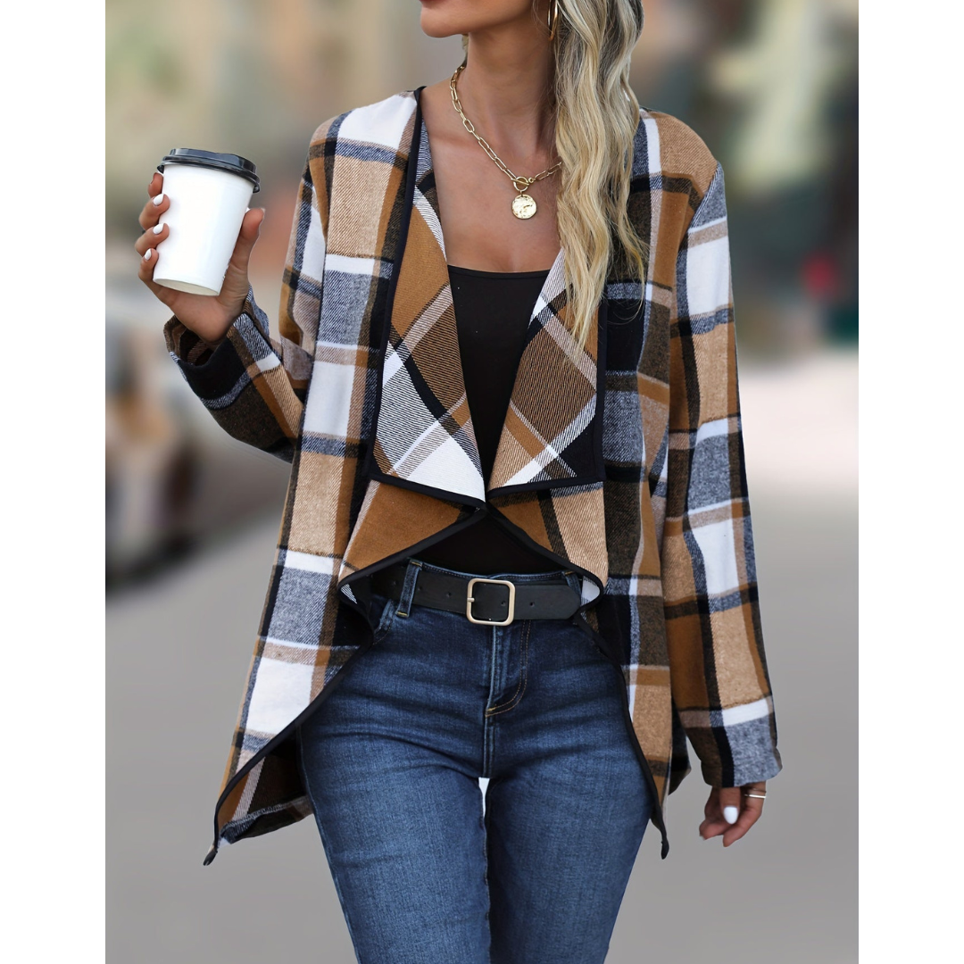 Donna con giacca a quadri marrone e bianca, top nero, jeans blu, cintura nera, collana dorata, tiene una tazza da caffè. Moda autunnale casual.
