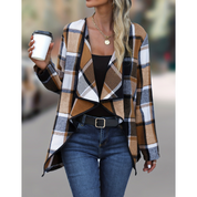 Donna con giacca a quadri marrone e bianca, top nero, jeans blu, cintura nera, collana dorata, tiene una tazza da caffè. Moda autunnale casual.