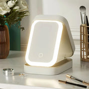 Specchio cosmetico LED rettangolare su tavolo da trucco, circondato da fiori bianchi e accessori di bellezza. Design moderno, illuminazione regolabile.