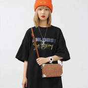 Giovane donna con borsa a tracolla marrone, indossa maglietta nera oversize e berretto arancione. Accessorio moda casual, stile urbano.