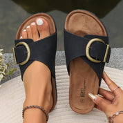 Sandali donna neri con fibbia dorata, suola in sughero, indossati su tappeto bianco. Scarpe estive eleganti, moda femminile, calzature comode.