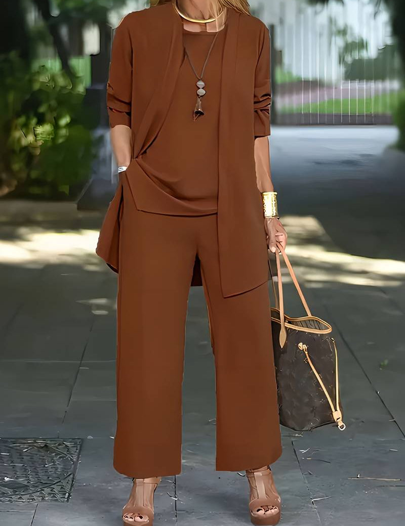 Donna con elegante completo marrone, pantaloni larghi e giacca lunga, indossa sandali con tacco e borsa di lusso. Look moda autunnale chic.
