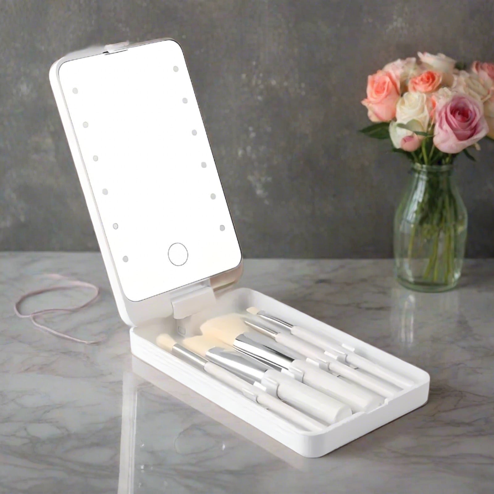Set di pennelli da trucco con specchio LED portatile su tavolo di marmo, accanto a vaso di fiori. Accessori bellezza, trucco professionale.