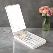 Set di pennelli da trucco con specchio LED portatile su tavolo di marmo, accanto a vaso di fiori. Accessori bellezza, trucco professionale.