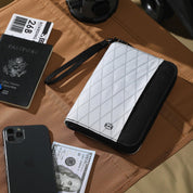 Portafoglio da viaggio bianco e nero con chiusura a zip, posizionato su un tavolo marrone, accanto a passaporto, smartphone e banconota da 100 dollari.