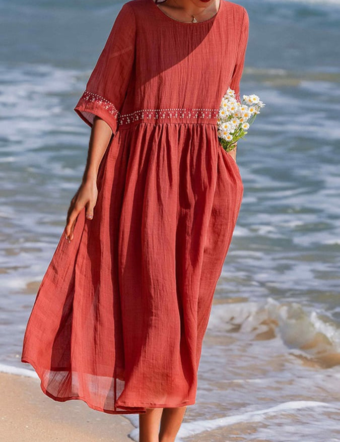 Donna in abito lungo rosso estivo, decorato con ricami, cammina sulla spiaggia con fiori bianchi in mano. Moda mare, abbigliamento donna elegante.