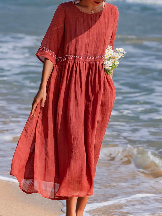 Donna in abito lungo rosso estivo, decorato con ricami, cammina sulla spiaggia con fiori bianchi in mano. Moda mare, abbigliamento donna elegante.