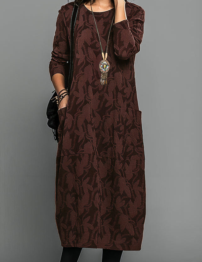 Abito lungo marrone con motivo floreale, maniche lunghe, tasche laterali, collana pendente. Elegante e casual, perfetto per l'autunno.