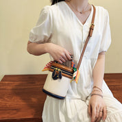 Donna con abito bianco tiene borsa a tracolla beige con dettagli in pelle marrone e foulard colorato. Accessorio moda elegante e chic.