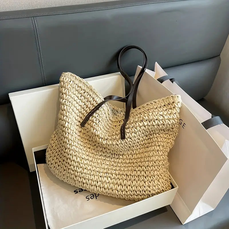 Borsa in paglia intrecciata con manici in pelle nera, posizionata su divano grigio, accanto a scatola e borsa shopping. Accessorio moda estivo elegante.