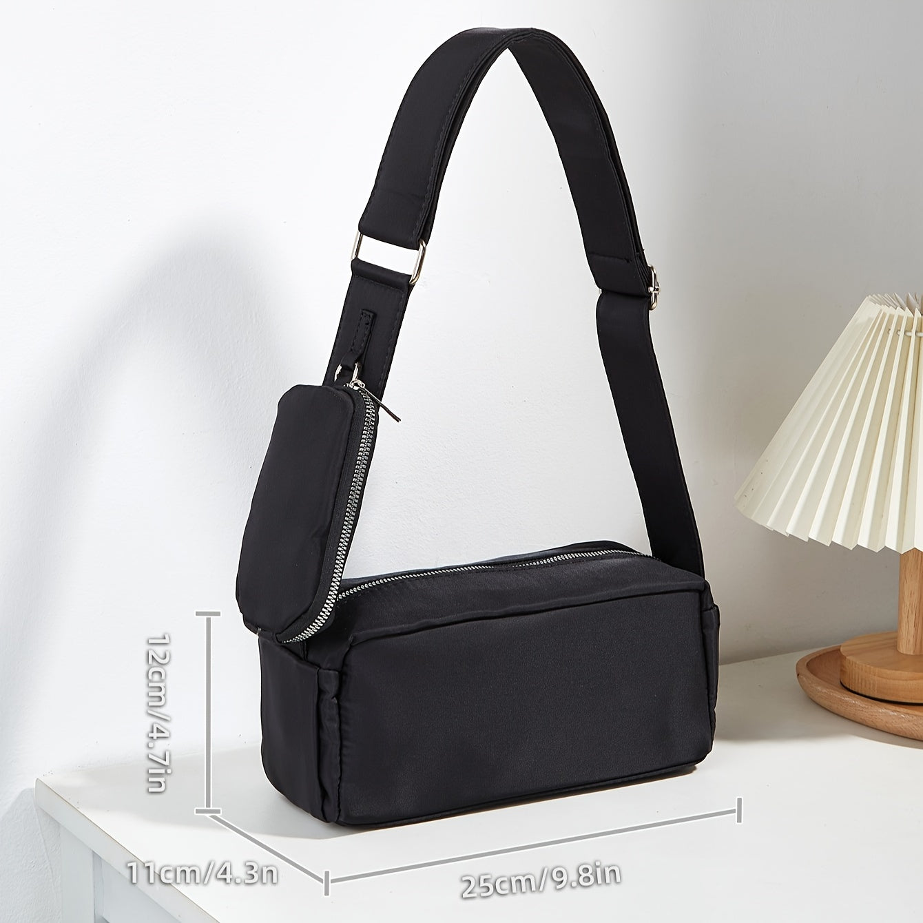 Borsa a tracolla nera unisex con tasca frontale con zip, dimensioni 25x11x12 cm, su tavolo bianco accanto a lampada. Accessorio moda casual.