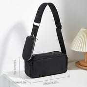 Borsa a tracolla nera unisex con tasca frontale con zip, dimensioni 25x11x12 cm, su tavolo bianco accanto a lampada. Accessorio moda casual.