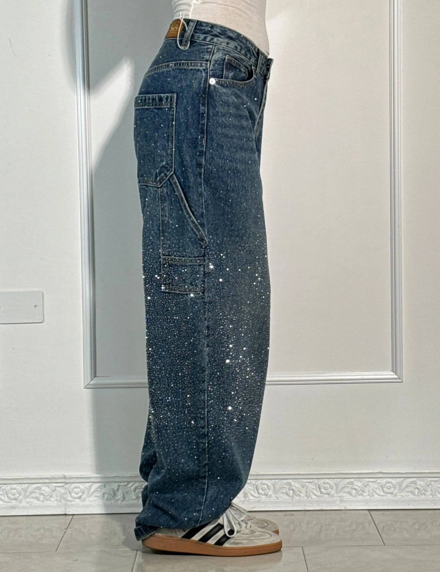 Jeans larghi in denim blu con paillettes scintillanti, tasche cargo, indossati con sneakers bianche e nere. Moda casual, stile urbano.