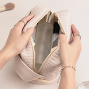 Borsa cosmetica beige trapuntata aperta, contenente flaconi di prodotti per la cura della pelle. Mani femminili con anelli e bracciale.