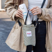 Donna con giacca beige tiene una borraccia bianca in una borsa a tracolla grigia. Accessorio pratico per bevande, ideale per viaggi e attività all'aperto.