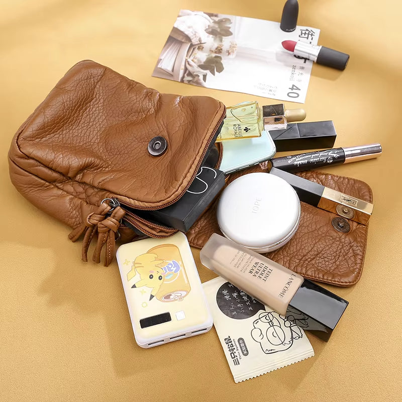 Borsa in pelle marrone con trucchi: rossetti, fondotinta, cipria, profumo, crema, power bank. Accessori moda e bellezza su sfondo beige.
