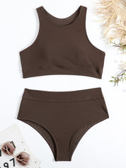 Costume da bagno donna marrone scuro a due pezzi, top sportivo e slip a vita alta, su sfondo bianco con occhiali da sole e decorazioni naturali.