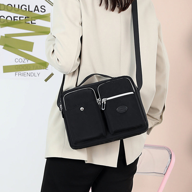 Borsa a tracolla nera unisex, design moderno, doppia tasca frontale con zip, indossata su giacca beige. Accessorio moda versatile e pratico.