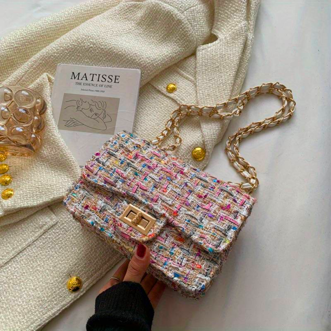 Borsa a tracolla in tweed multicolore con catena dorata su giacca bianca, accanto a libro d'arte e bicchiere. Elegante accessorio moda femminile.