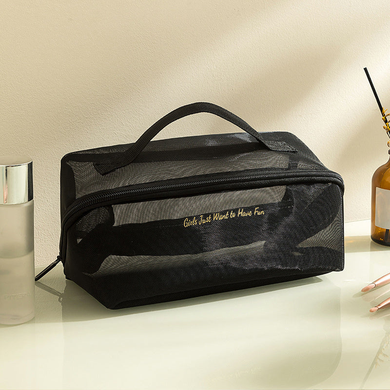 Borsa cosmetica nera in rete con manico, scritta dorata "Girls Just Want to Have Fun", su tavolo con flacone trasparente e bottiglia marrone.