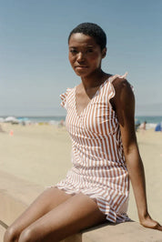 Donna in abito estivo a righe bianche e marroni, seduta su un muretto in spiaggia. Moda mare, abbigliamento estivo, look casual elegante.