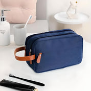 Borsa da toilette blu con manico in pelle marrone su tavolo bianco, accanto a spazzolino e dispenser sapone. Accessorio da viaggio elegante e pratico.