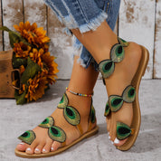Sandali estivi da donna con decorazioni a forma di piuma di pavone verde, suola piatta, indossati con jeans strappati. Accessorio moda casual.