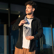 Giovane uomo con marsupio grigio, camicia nera e pantaloni marroni, in piedi all'aperto. Accessorio moda uomo, stile casual urbano.