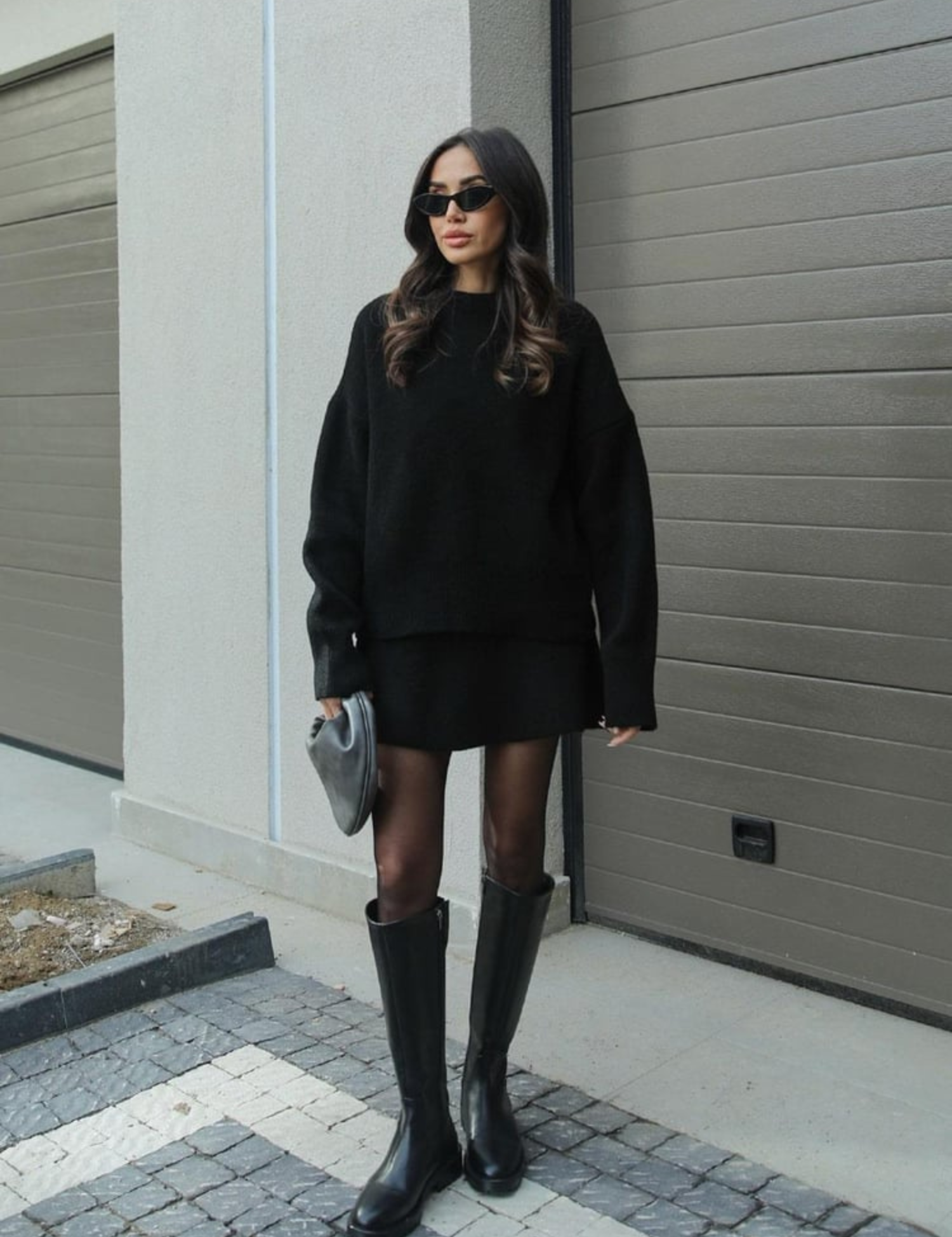 Donna con occhiali da sole indossa maglione nero oversize, minigonna e stivali alti neri. Look moda autunnale elegante e casual.
