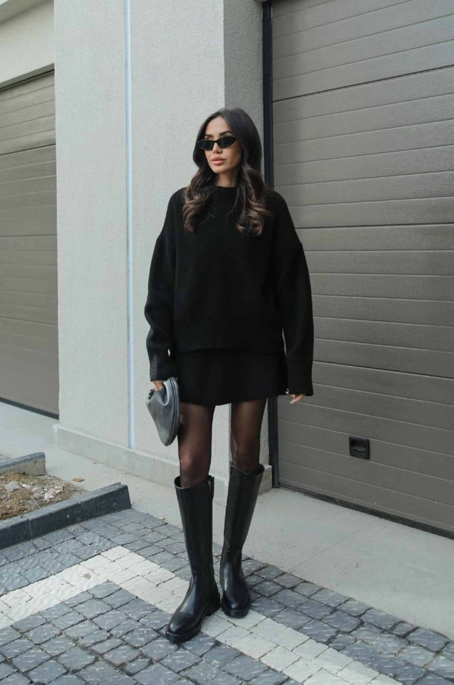 Donna con occhiali da sole indossa maglione nero oversize, minigonna e stivali alti neri. Look moda autunnale elegante e casual.