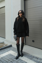 Donna con occhiali da sole indossa maglione nero oversize, minigonna e stivali alti neri. Look moda autunnale elegante e casual.