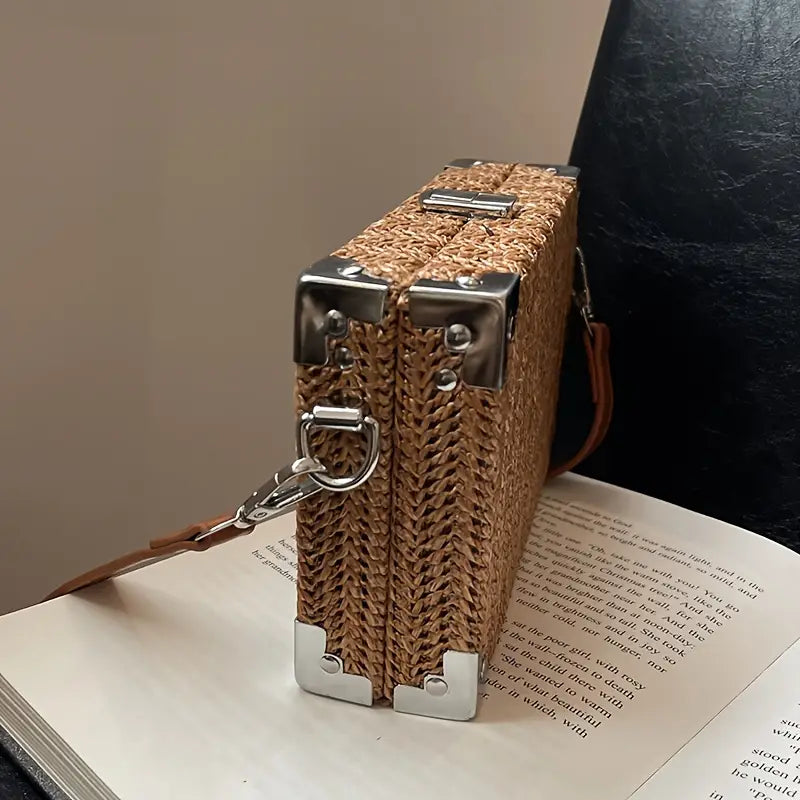 Borsa a tracolla in paglia intrecciata con dettagli in metallo argentato, design rettangolare, su sfondo di libro aperto. Elegante e moderna.