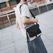 Uomo con borsa a tracolla nera, indossa maglietta grigia e jeans, passeggia all'aperto. Accessorio moda casual, stile urbano.