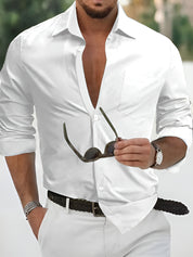 Camicia bianca elegante da uomo, maniche lunghe, abbinata a pantaloni bianchi e cintura intrecciata. Accessorio: occhiali da sole. Look estivo sofisticato.