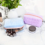 Due astucci da trucco in velluto, uno azzurro e uno viola, con cerniera, su un tavolo di marmo, accanto a pennelli e cosmetici, vaso di fiori sullo sfondo.